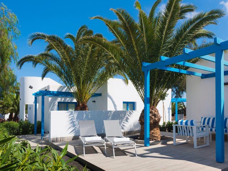 Terraza con pérgolas azules, edificios blancos y tumbonas rodeadas de palmeras y vegetación.