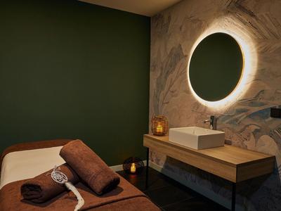 Gemütlicher Behandlungsraum mit Massageliege, dunkler Wand und rundem, beleuchtetem Spiegel.