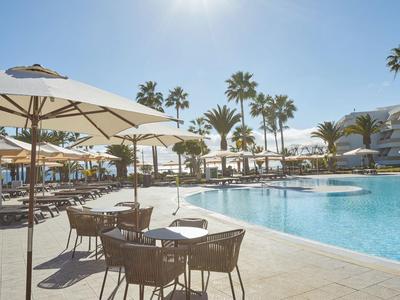 Hotelpool mit Sonnenschirmen, Liegestühlen und Tischen bei sonnigem Wetter und Palmen.