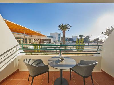 Balkon mit zwei Stühlen und Tisch mit Blick auf Pool und Palmen bei klarem Himmel.