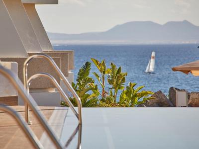 Infinity-Pool mit Metallgeländer, grünen Pflanzen und Segelboot auf ruhigem blauem Meer im Hintergrund.