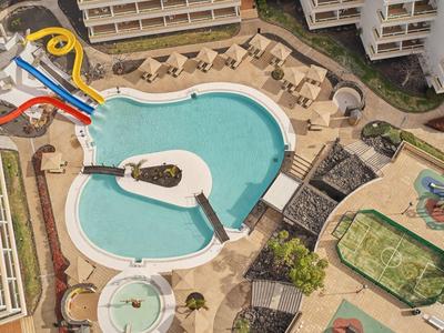 Großer blauer Pool mit Wasserrutschen, Liegen und umliegenden Gebäuden aus der Vogelperspektive.