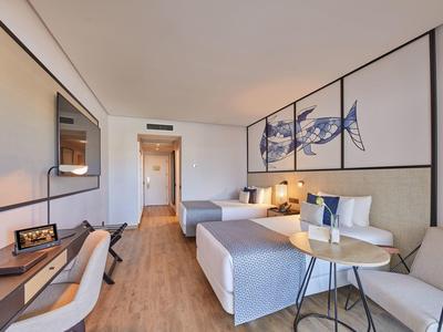 Modernes Hotelzimmer mit zwei Betten, Schreibtisch, Sofa und Fischwandbild in hellem Holzdekor.