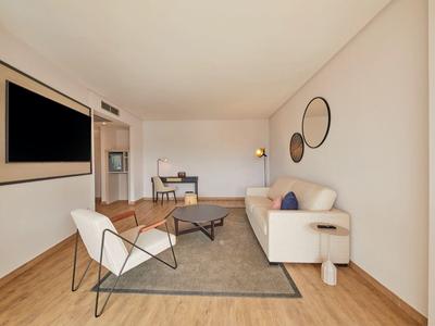 Helles Wohnzimmer mit weißem Sofa, zwei Sesseln, grauem Teppich und großem Fernseher an der Wand.