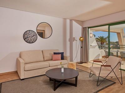 Helles Wohnzimmer mit beige Sofa, zwei runden Spiegeln, braunem Tisch und Stuhl vor Balkon mit Meerblick.