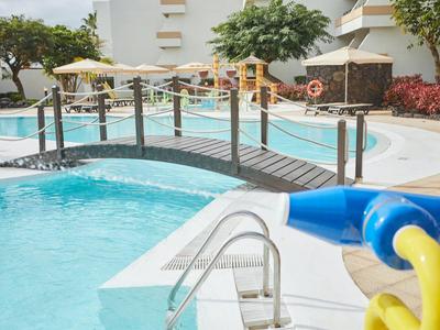 Schwimmbad mit Brücke, Wasserrutsche und Sonnenschirmen in hellem, sonnigem Umfeld.