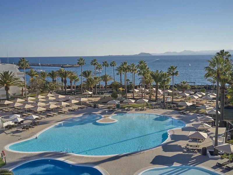 Großer Pool mit Liegestühlen und Palmen, dahinter Meer und klarer blauer Himmel am Morgen.