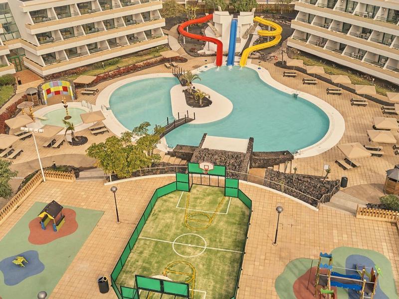 Hotelanlage mit Pool, Wasserrutschen, Liegestühlen und einem Spielplatz im Vordergrund.