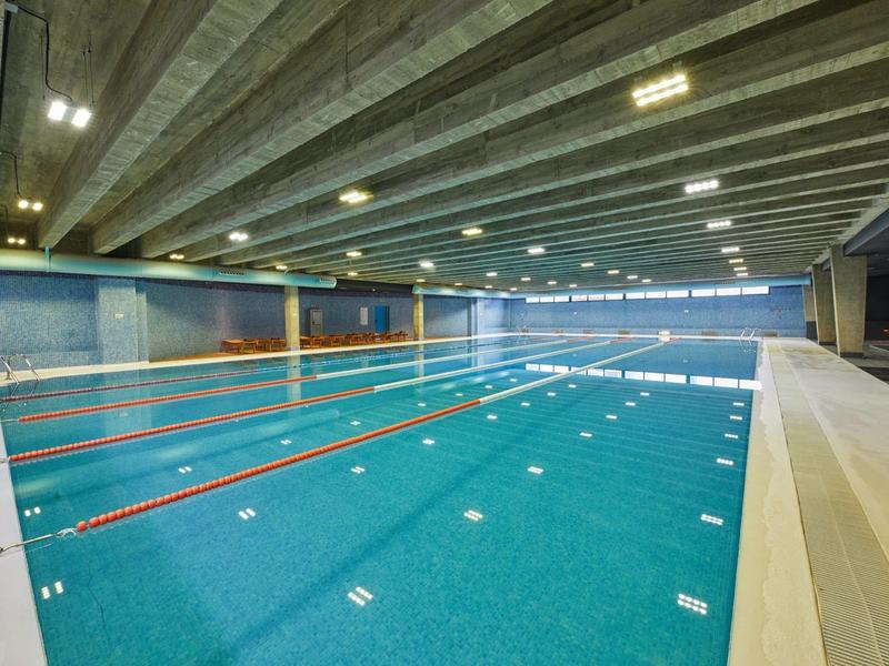 Hallenbad mit mehreren Schwimmbahnen, grauen Deckenbalken und blauen Wänden.