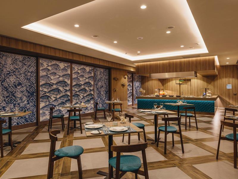 Moderner Gastraum mit Holzwänden, blau-grünen Stühlen und beleuchtetem Tresen in warmem Licht.