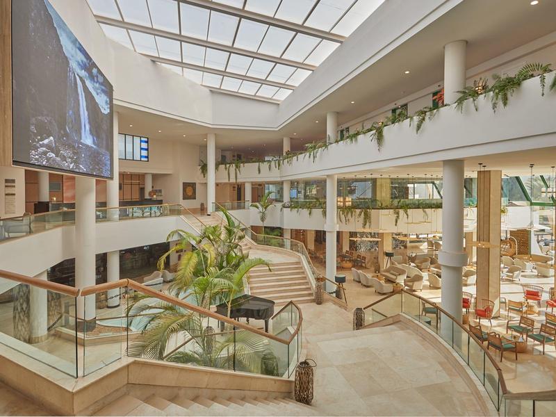 Helles modernes Einkaufszentrum mit Rolltreppen, Glasgeländern und großen Pflanzen.