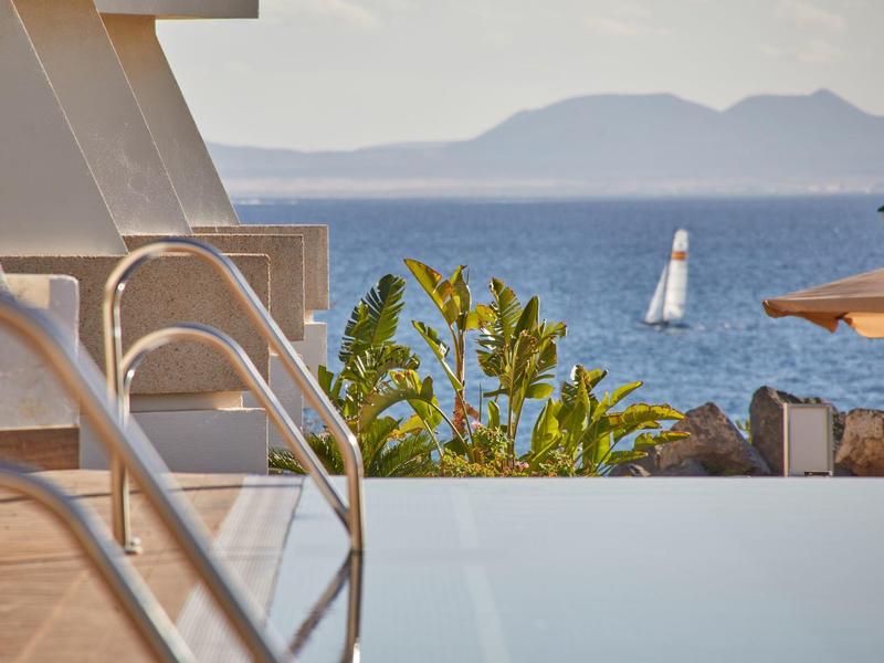 Infinity-Pool mit Metallgeländer, grünen Pflanzen und Segelboot auf ruhigem blauem Meer im Hintergrund.