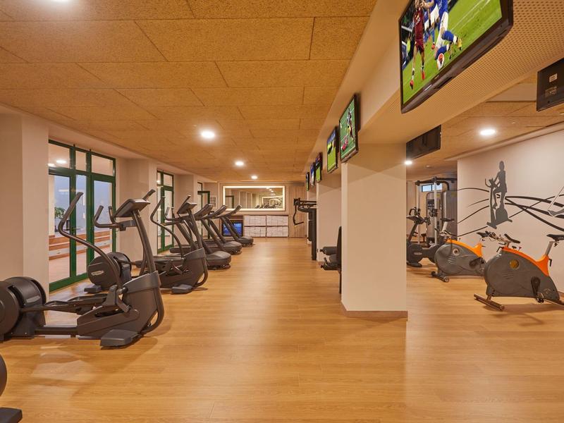 Moderner Fitnessraum mit Holzfußboden, Cardio-Geräten und TV-Bildschirmen an der Decke.