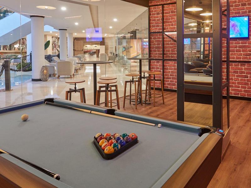 Moderne Lounge mit grauem Billardtisch, braunem Holzfußboden und offenem Barbereich im Hintergrund.