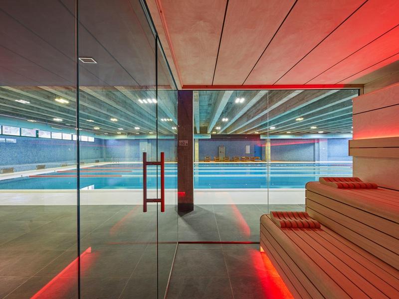 Moderner Wellnessbereich mit Sauna aus Holz und großem Schwimmbecken hinter Glaswand.