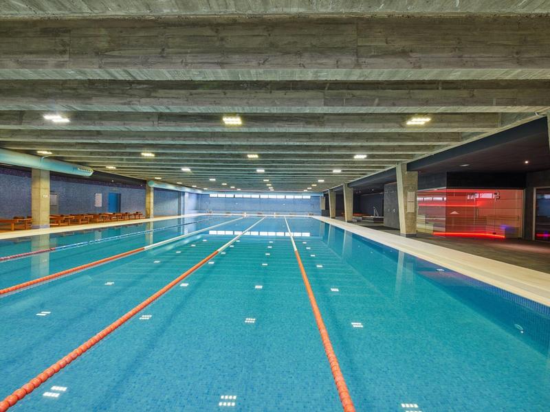 Schwimmbecken mit sechs Bahnen in moderner Halle, blaues Wasser, weiße und rote Linien.