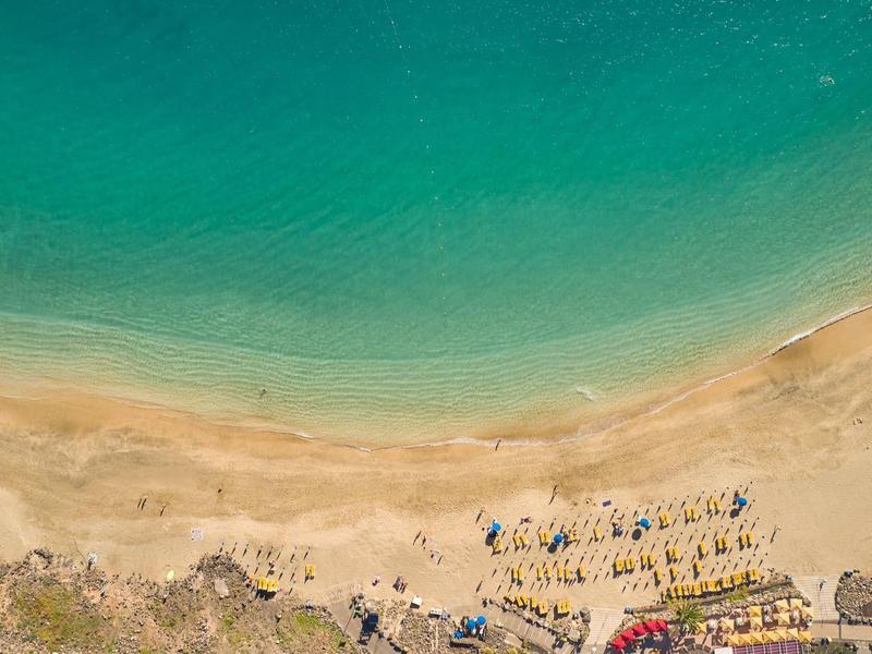 Luftaufnahme von Strand mit türkisblauem Wasser, Sand, Sonnenschirmen und Liegestühlen.