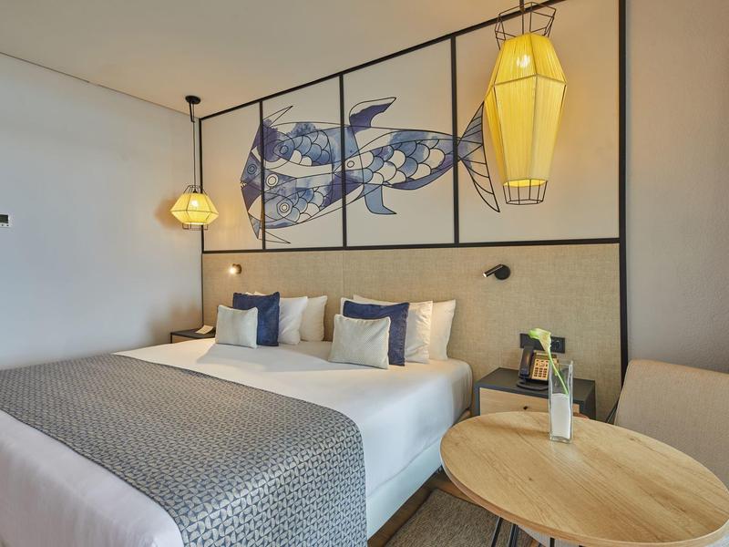 Modern eingerichtetes Hotelzimmer mit Doppelbett, drei Kissen, rundem Tisch und dreiteiligem Wandbild.