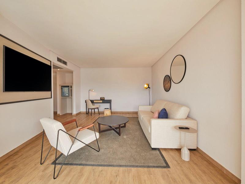 Helles Wohnzimmer mit weißem Sofa, zwei Sesseln, grauem Teppich und großem Fernseher an der Wand.