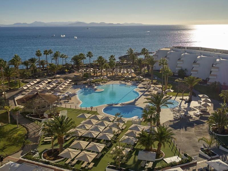 Luxuriöse Hotelanlage mit Pool, grünen Palmen und Blick auf das helle Meer bei Sonnenuntergang.