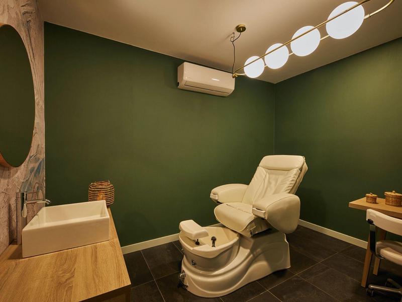 Behandlungsraum mit grünen Wänden, weißem Liegestuhl, Waschbecken, Holzspiegel und modernem Licht.