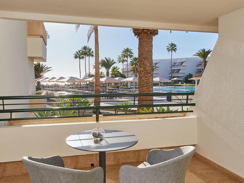 Balkon mit kleinem runden Tisch, zwei Stühlen, Blick auf Palmen und Hotelgebäude bei Sonnenschein.