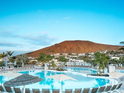 Runder Pool mit klar blauem Wasser vor einem roten Vulkan unter blauem Himmel.