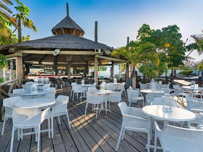 Freiluftrestaurant auf Holzterrasse mit weißen Tischen und Stühlen, umgeben von Palmen.