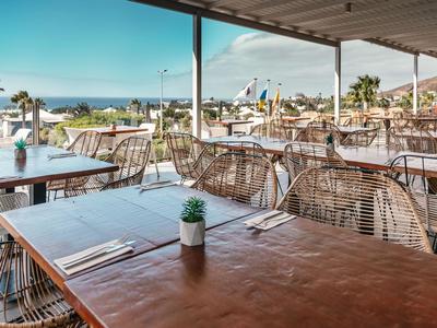 Überdachte Terrasse eines Restaurants mit Tischen, Stühlen und Blick auf Palmen und das Meer.