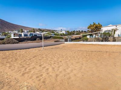 Großer, leerer Volleyballplatz mit Sand und zwei Pfosten vor weißen Gebäuden und Bergen.