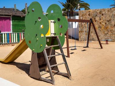 Spielplatz mit Sand, grünem Baumrutschen-Klettergerüst und Schaukel vor buntem Gebäude.