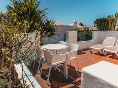 Eine sonnige Terrasse mit weißen Stühlen, Tisch und Sonnenliegen, umgeben von Pflanzen.