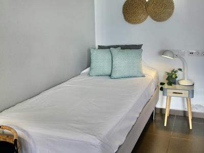 Schlichtes Einzelbett mit weißen Laken, grauen und blauen Kissen, kleinem Nachttisch und Wanddeko aus Flechtmaterial.