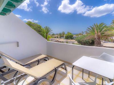 Balkon mit zwei Liegestühlen, kleinem weißen Tisch, weißer Wand und Blick auf Palmen unter blauem Himmel.