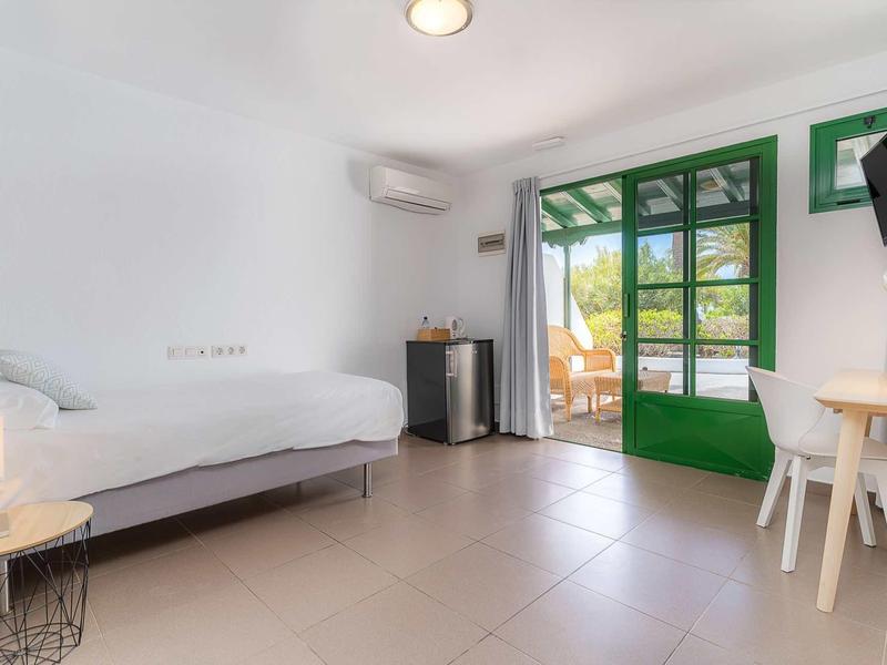 Helles Zimmer mit Einzelbett, grünem französischem Fenster und einfachem Holztisch mit Stuhl.