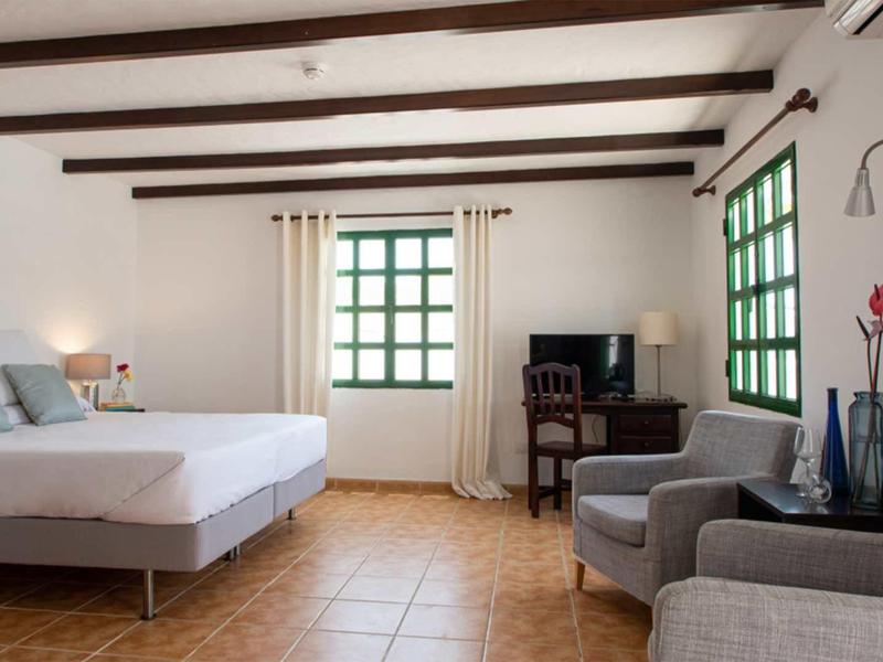 Helles Schlafzimmer mit Holzbalken, zwei Sesseln, TV, großen Fenstern und Terrakottaboden.