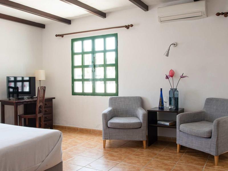 Helles Zimmer mit zwei grauen Sesseln, grünem Fenster, Fernseher und Bett mit Holzboden.