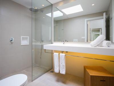 Helles Badezimmer mit Glas-Dusche, großem Spiegel, weißen Handtüchern und Holzhocker unter Waschbecken.