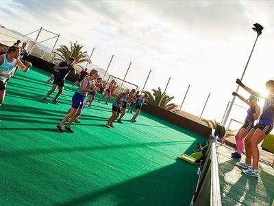 Menschen machen Sport auf grünem Spielfeld, zwei führen Übung vor bei sonnigem Himmel.