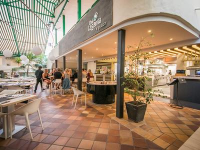 Großes, helles Restaurant mit braunen Kacheln, weißen Tischen und grünen Pflanzen, viele Gäste sitzen.