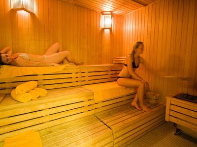 Zwei Frauen entspannen in einer holzverkleideten Sauna mit warmem Licht und gelben Handtüchern.