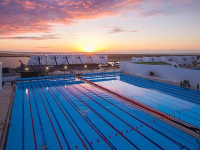 Großes Schwimmbecken mit Bahnen bei Sonnenuntergang, klare Wasseroberfläche, bunte Wolken am Himmel.