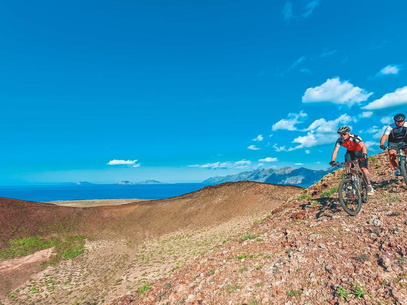 Two cyclists on a rocky mountain ridge under a clear blue sky.