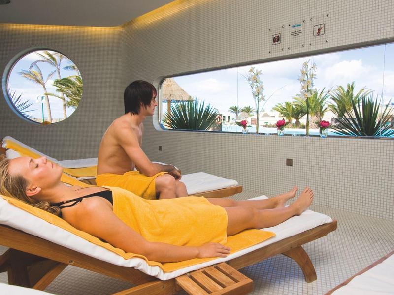 Two people relaxing on loungers in a modern spa with large window and plants.