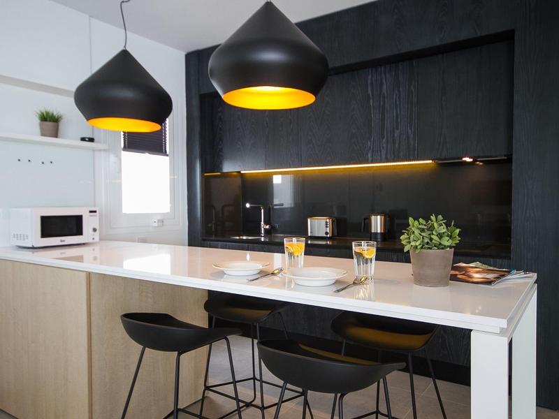 Modern hotel kitchen with wooden countertop, black stools, and two pendant lights.