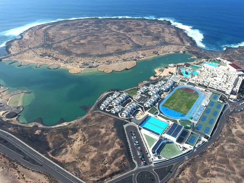 Aerial view of a coastal island with a harbor, sports facilities, surrounded by water and sea.