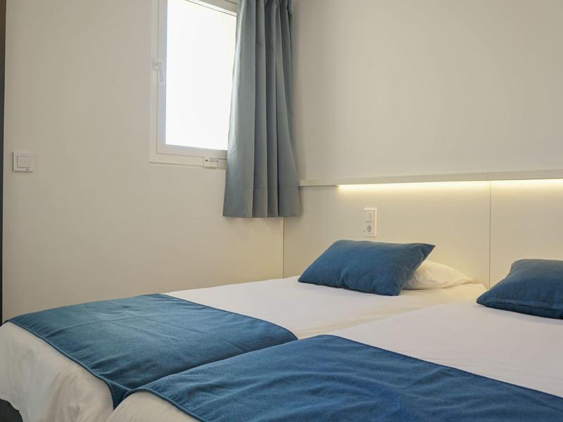 Bright hotel room with two single beds, blue pillows, and blankets next to a window with curtain.