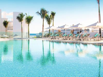 Grande piscine avec chaises longues et palmiers dans une station ensoleillée près de la mer.