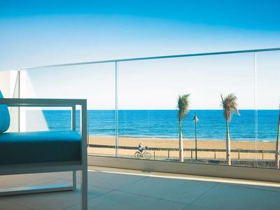 Blick auf das Meer von einer Terrasse mit Glasgeländer, blauer Stuhl links und Palmen am Strand.