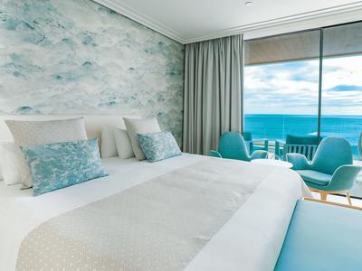 Helles Schlafzimmer mit großem Bett, blau-weißer Tapete und Meerblick durch bodentiefe Fenster.